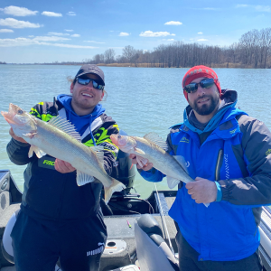 Two men stand on a boat in the middle of Lake Erie. Each of them holds up the walleye they caught. They look proud of their catch.